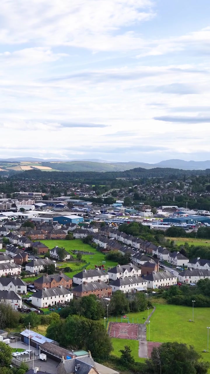Drone svever over forstedene i Dundee, avslører hustak, grøntområder og fjerne åser i dagslys.