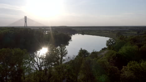 Aerial-shot-of-a-wide-calm-river-with-a-large-cable-bridge-visible-in-the-distance