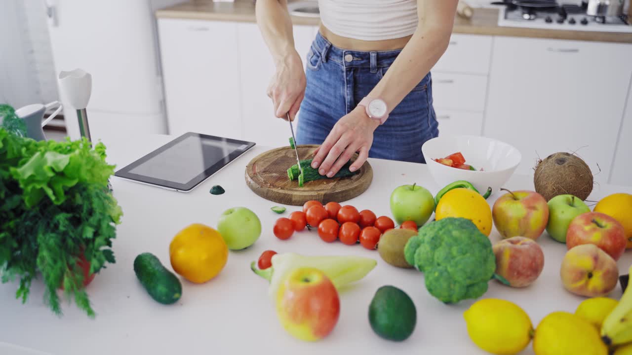 Delicious fruit and vegetables on a table and woman cooking. Housewife is cutting green cucumbers on a wooden board for making fresh salad in the kitchen.