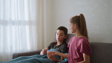 sister hands over thermometer to sick brother seated on couch, as he prepares to check his temperature by placing it under his arm, natural light filters through window