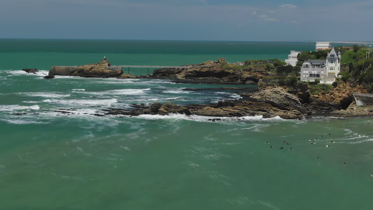 Rocher de la Vierge and Villa Belza in Biarritz, surfers ride waves near rocky coast, Basque Country, France. Aerial forward, copy space