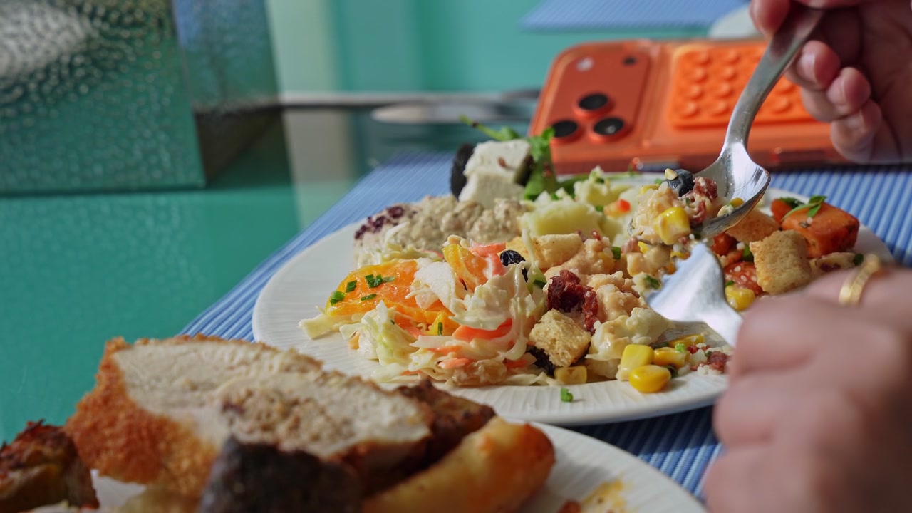 Primer plano de la mano de una persona usando una cuchara para comer una ensalada fresca y saludable con maíz y picatostes. La comida servida está sobre un mantel individual azul en una mesa de comedor interior con un teléfono naranja de fondo.