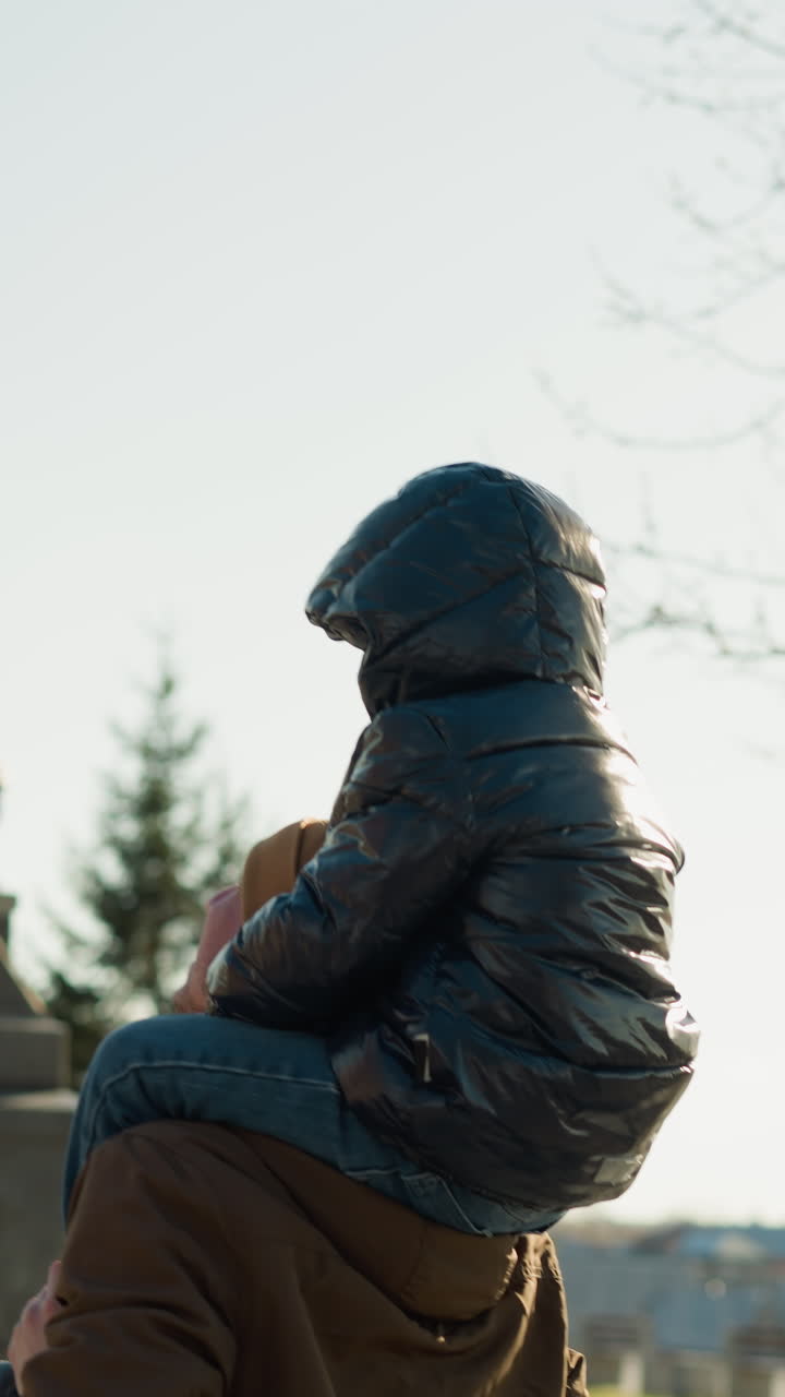 un primer plano de un padre llevando a su hijo en sus hombros juguetón, el padre lleva una chaqueta marrón, y el hijo lleva unachaqueta negra, con una vista borrosa del paisaje urbano