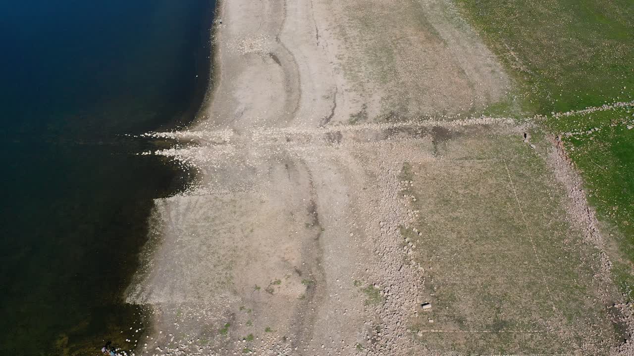 vuelo con un dron a lo largo de la orilla de un embalse donde vemos el agua, la tierra blanquecina con restos de piedras que forman estructuras con un camino sumergido y tierra con plantas verdes invierno en españa