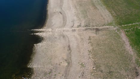 flight-with-a-drone-along-the-shore-of-a-reservoir-where-we-see-the-water,-the-whitish-land-with-remains-of-stones-forming-structures-with-a-submerged-path-and-land-with-green-plants-winter-in-Spain