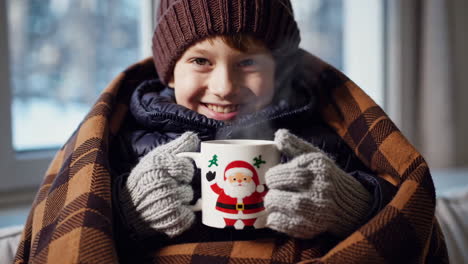 young boy bundled in a blanket holding a steaming christmas mug in winter