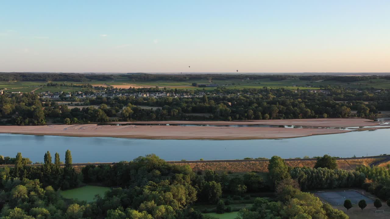 Aerial rises over bucolic town by quiet river, distant hot air balloons