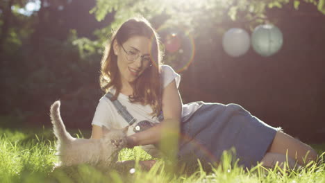 Young-Caucasian-woman-in-glasses-lying-on-the-grass-and-petting-kitty-cat-in-the-park