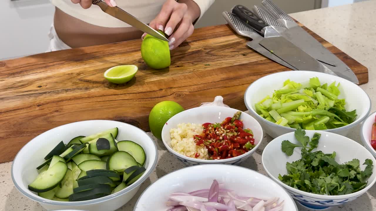A person slices a lime on a wooden cutting board surrounded by bowls of chopped cucumber, chili, herbs, and vegetables under bright kitchen lighting