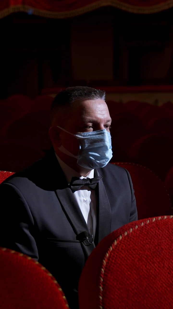 Man sits in the middle of empty theater hall with red velvet chairs. Actor is alone in theater. Lockdown and pandemic concept. Vertical video