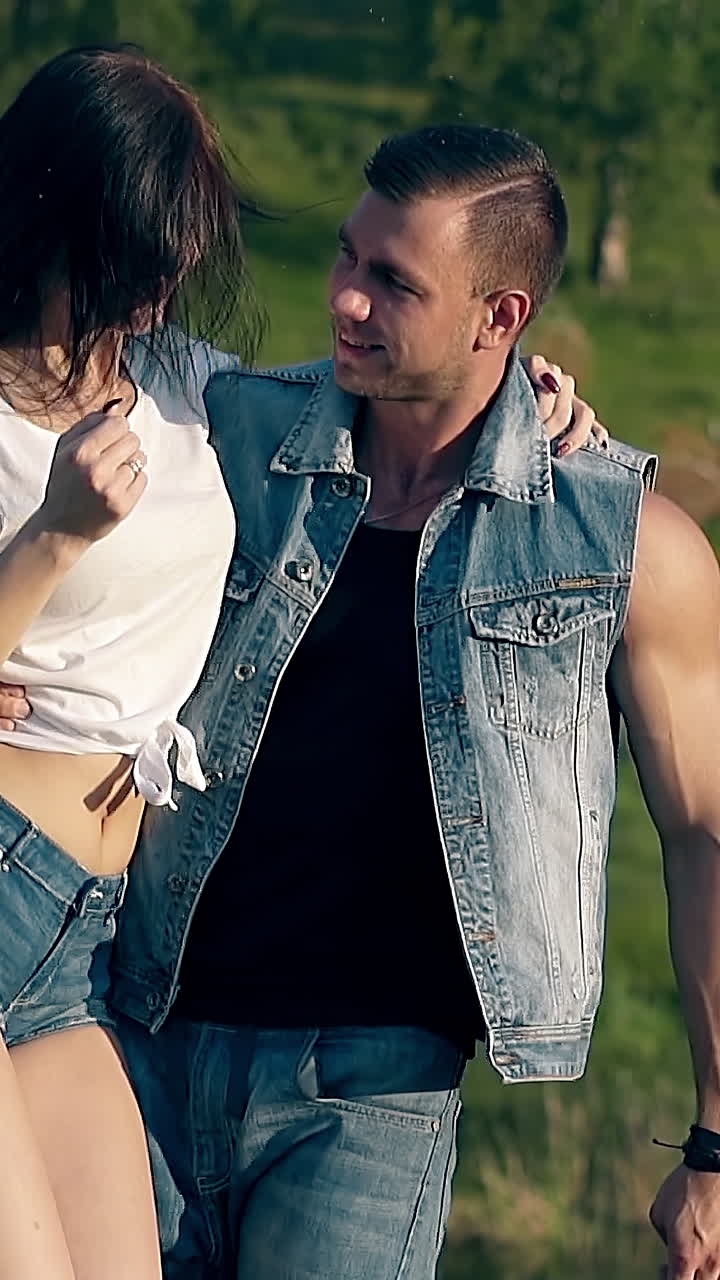 handsome man in denim shorts and vest holds pretty brunette lady in arm and spins against green grass slow motion