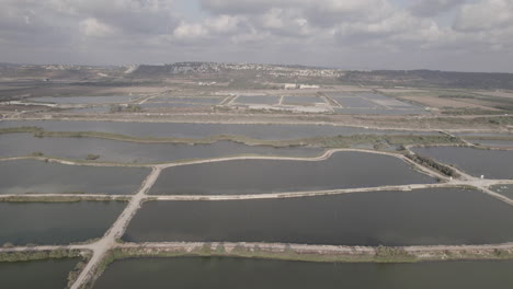 Vista-Aérea-De-Estanques-De-Peces-En-El-Borde-De-Un-Lago-Natural-Al-Pie-De-Las-Montañas,-Una-Vasta-Y-Verde-Reserva-Natural,-Vista-Amplia-Con-Una-Carretera-Principal-Al-Lado-De-Las-Piscinas,-Deslizándose-Hacia-La-Izquierda