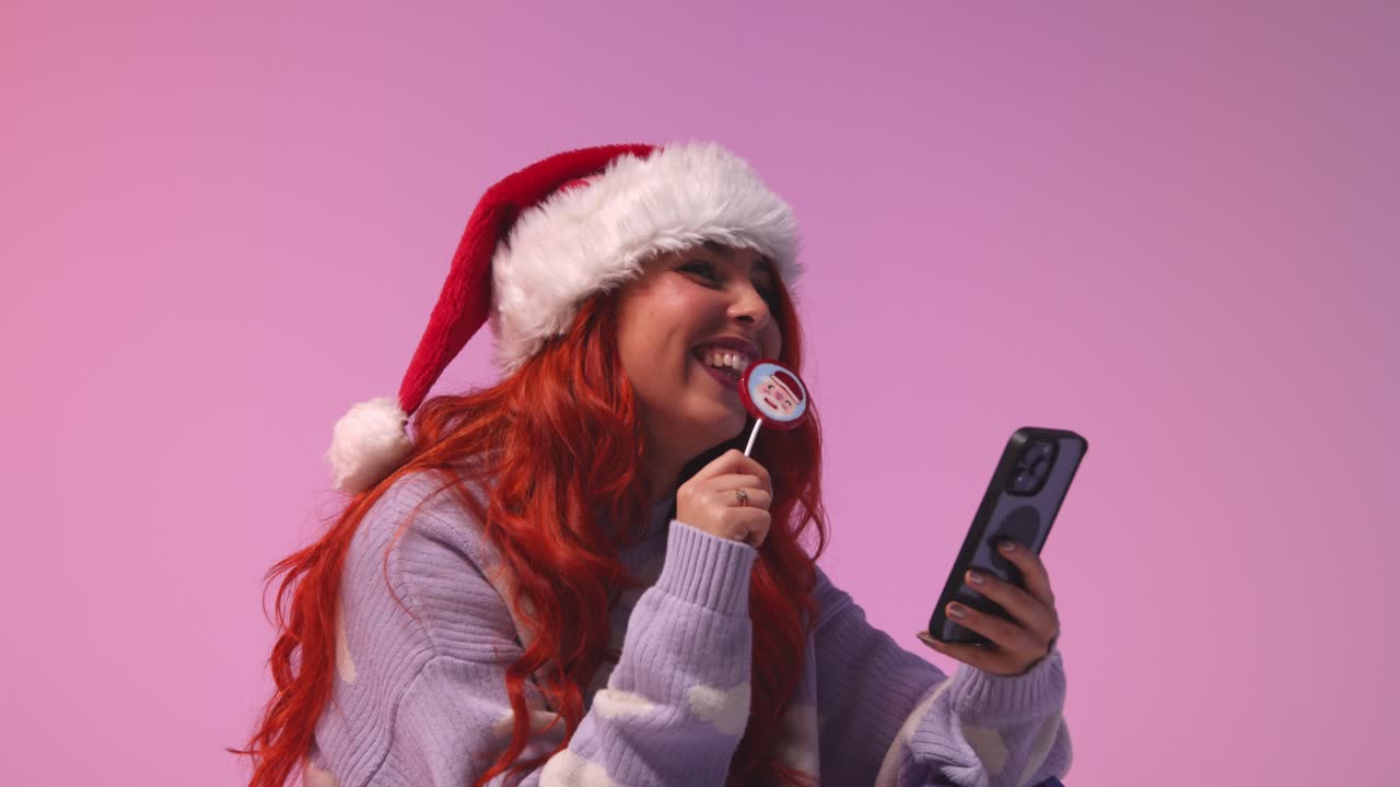 fotografía de estudio de una mujer joven de la generación z con sombrero de papá noel comiendo caramelos y mirando el teléfono móvil 1