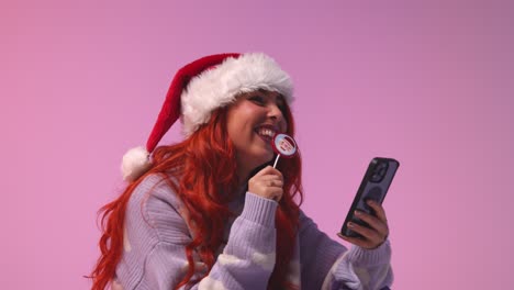 Studio-Shot-Of-Young-Gen-Z-Woman-Wearing-Christmas-Santa-Hat-Eating-Candy-Lollipop-Looking-At-Mobile-Phone-1