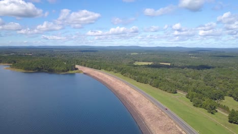 cars driving on a lake dam surrounded by forest and blue water and distant mountains