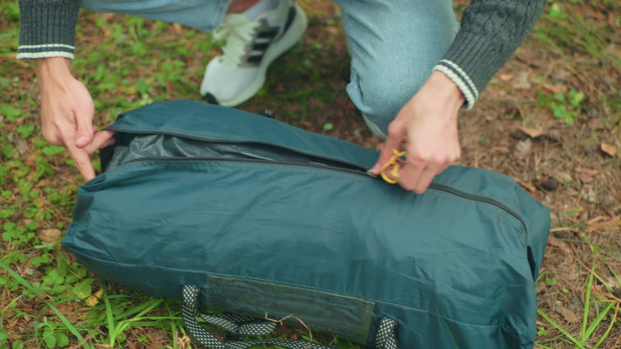 close up of student squatting on forest ground while pulling tent bag out of zipped carrier bag, surrounded by green grass and natural environment, preparing for outdoor camping setup