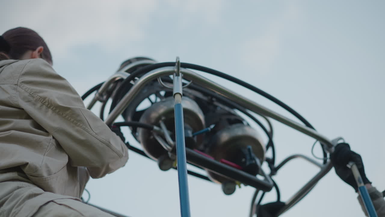 man assists woman with burner rig installation inside wicker basket during hot air balloon preflight setup by roadside under cloudy sky with red envelope and lush grassy field backdrop