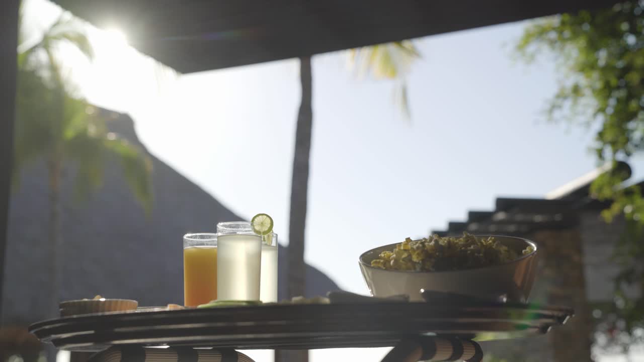 a waiter puts down a tray of snacks and drinks, including popcorn and orange juice in a tropical resort environment while the sun is beaming in the background