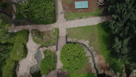 Drone-flies-over-a-beautiful-garden-and-rises-upwards-while-a-bird-passes-below-it,-at-the-ground-there-is-a-small-water-channel-with-a-fish-pond