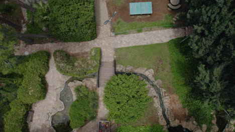 drone vuela sobre un hermoso jardín y se eleva mientras un pájaro pasa por debajo, en el suelo hay un pequeño canal de agua con un estanque de peces