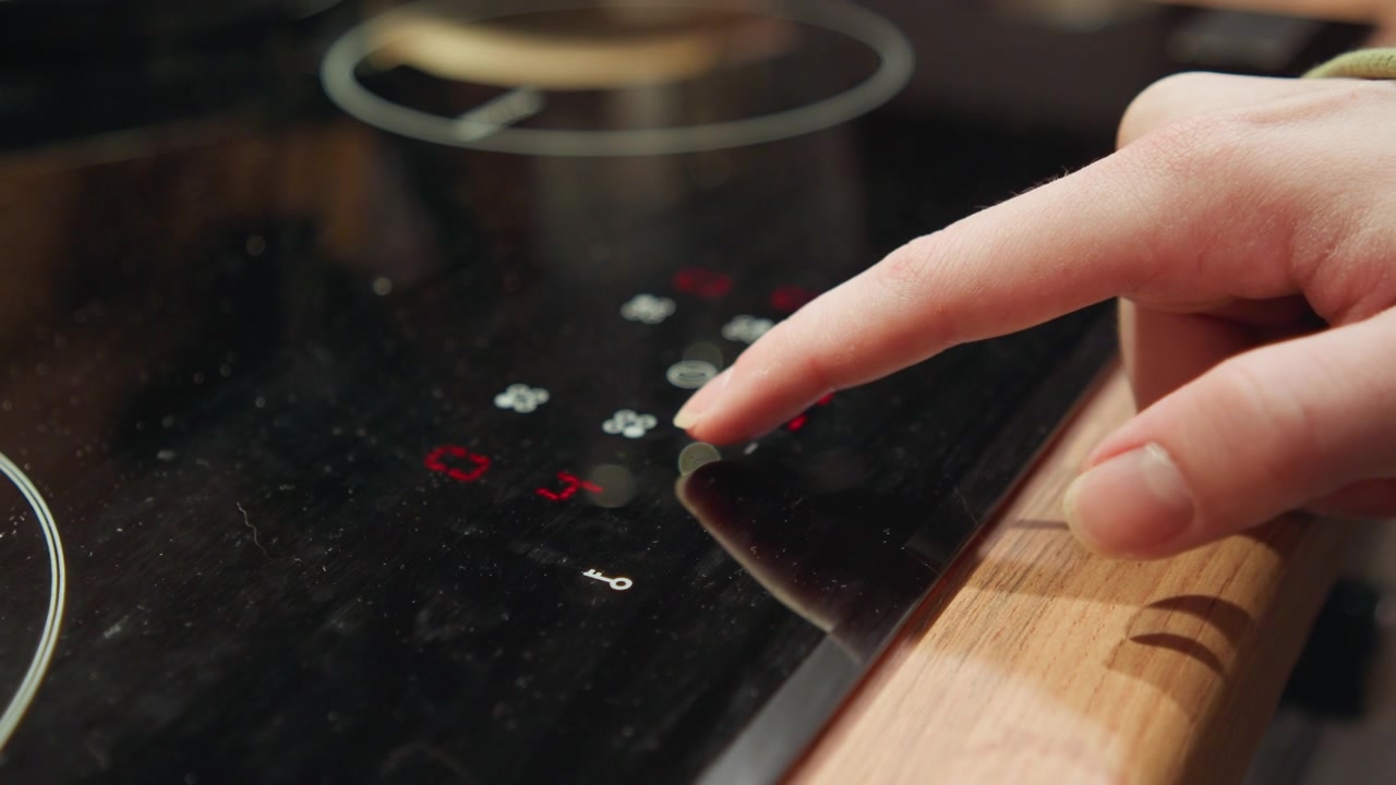 Close-up of fingertip pressing glowing red power control on glossy black induction cooktop, with nearby touch symbols lit, visible downward motion, slight pressure, and reflective surface detail