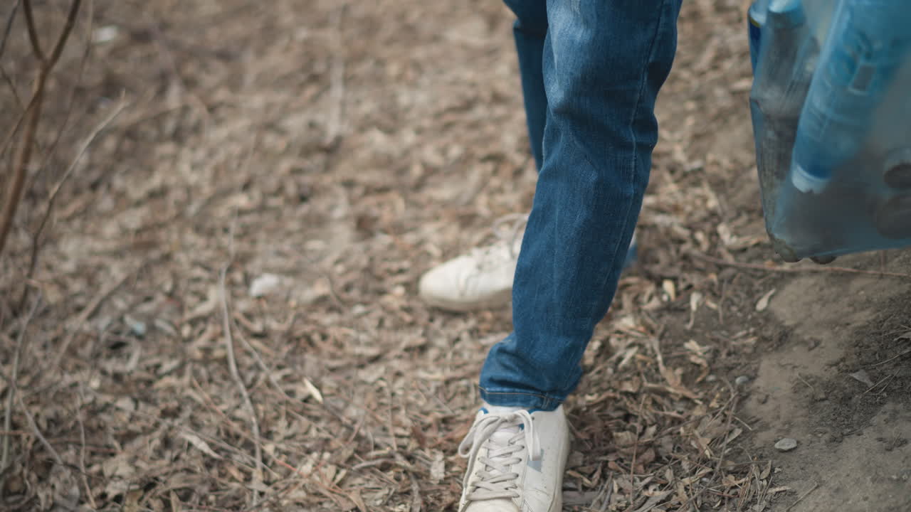 Close-up of person wearing white sneakers and blue jeans standing on dry leaf-covered ground during environmental cleanup, holding blue plastic bag with collected litter