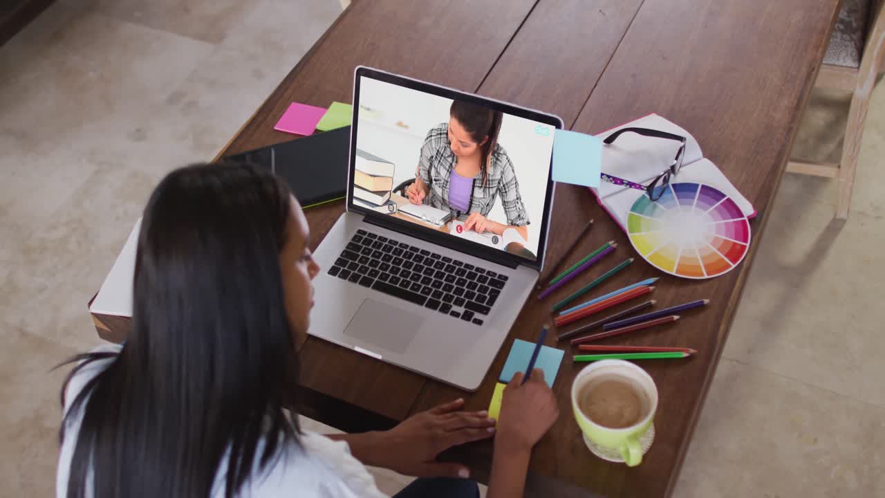 Profesora caucásica usando una laptop en videollamada con una estudiante, tomando apuntes.