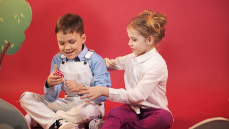 small boy and girl playing with red and white lollipop. cheerful friends sitting on the floor in red studio background.