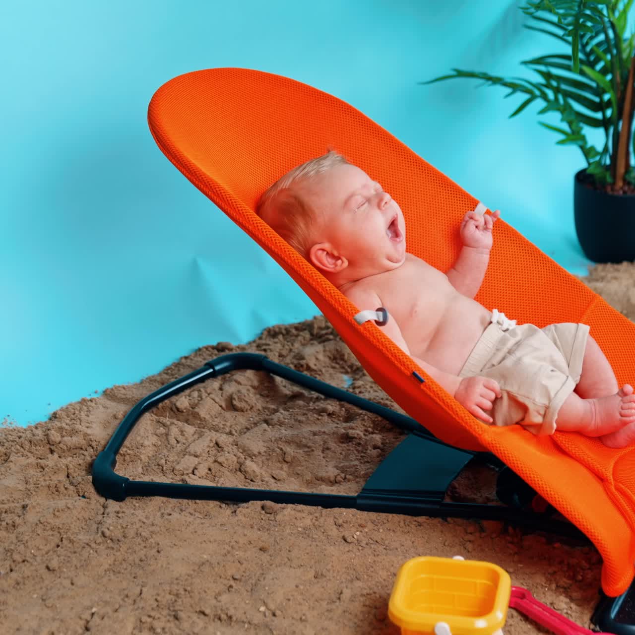 Adorable nouveau-né en short, bâillant et s’étirant dans une chaise orange. Bébé sur une plage improvisée en studio. Vue de côté