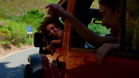 portrait of laughing female friends looking out of windows of open top car on road trip