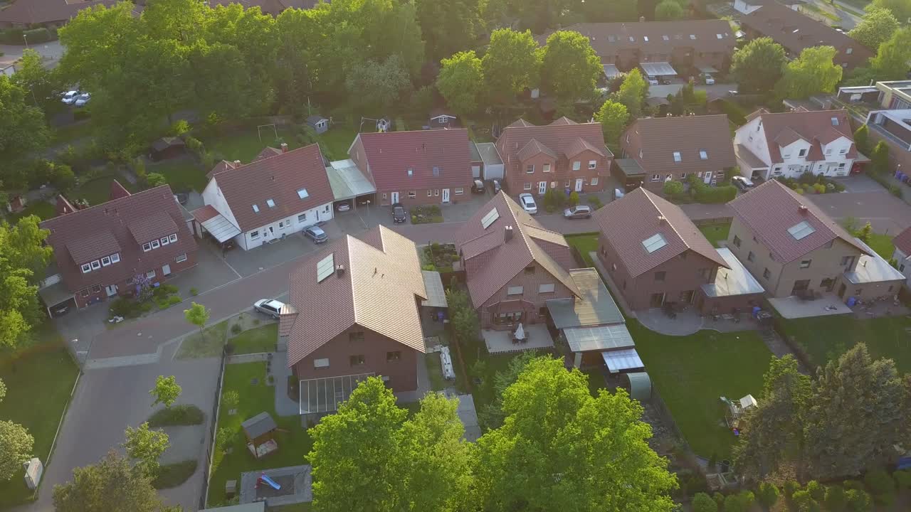 A drone circles a neighborhood in Gifhorn, Germany, showing green backyards with lush lawns and trees. Sunlight flares on the lens, adding warmth to the scenic, peaceful view.