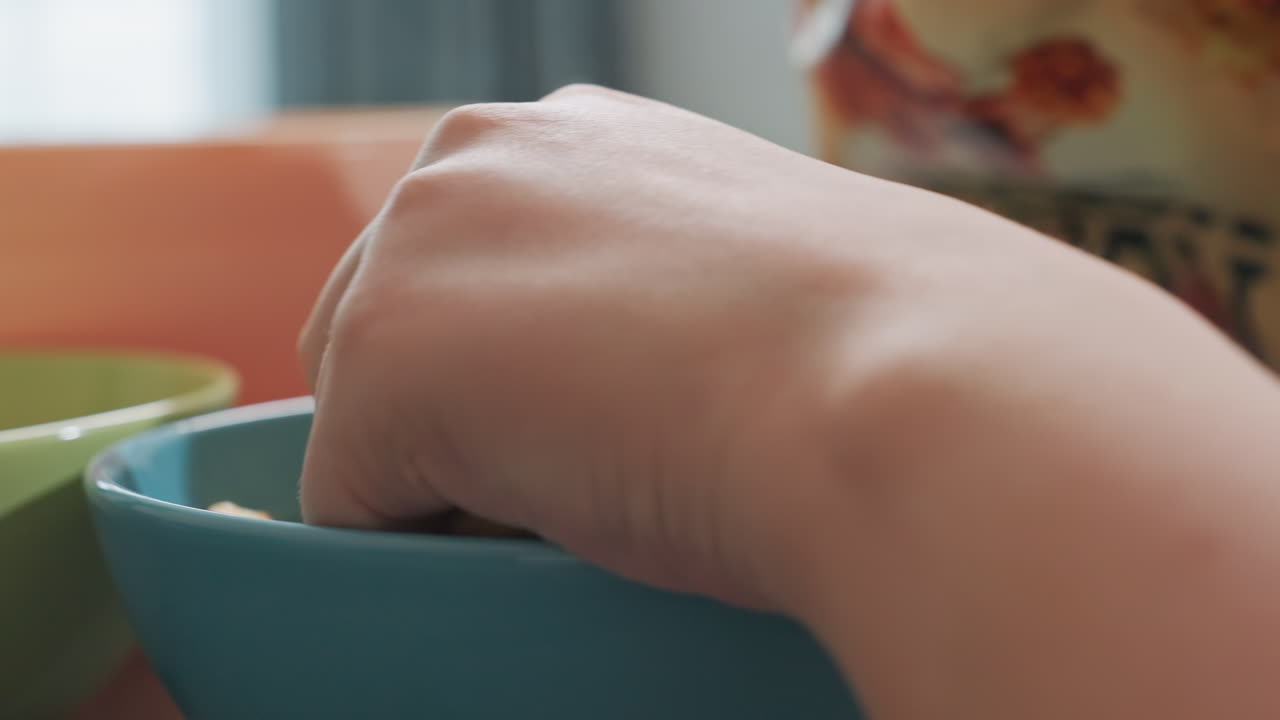 Close up hand view of person returning popcorn into packet on table with blue and green bowls on side and blurred person passing by in background, snack preparation with casual indoor setting