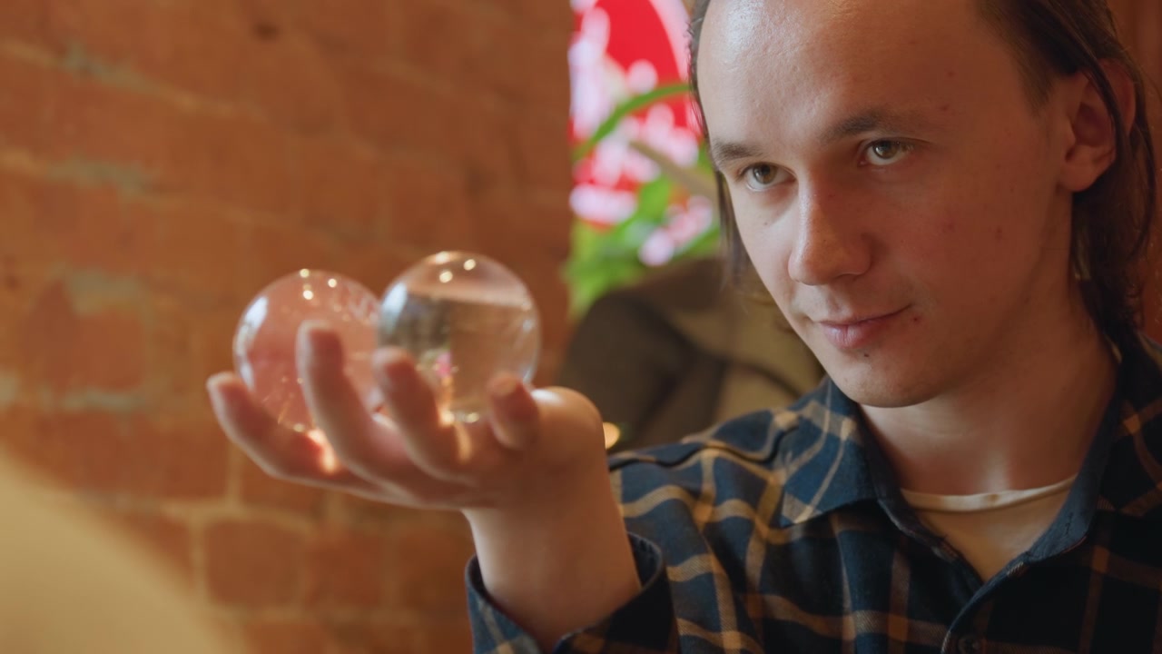Primer plano de un hombre sosteniendo esferas de cristal transparentes en su mano contra un fondo de pared de ladrillo interior. El sujeto mantiene un enfoque constante en los objetos con una iluminación suave que resalta su transparencia