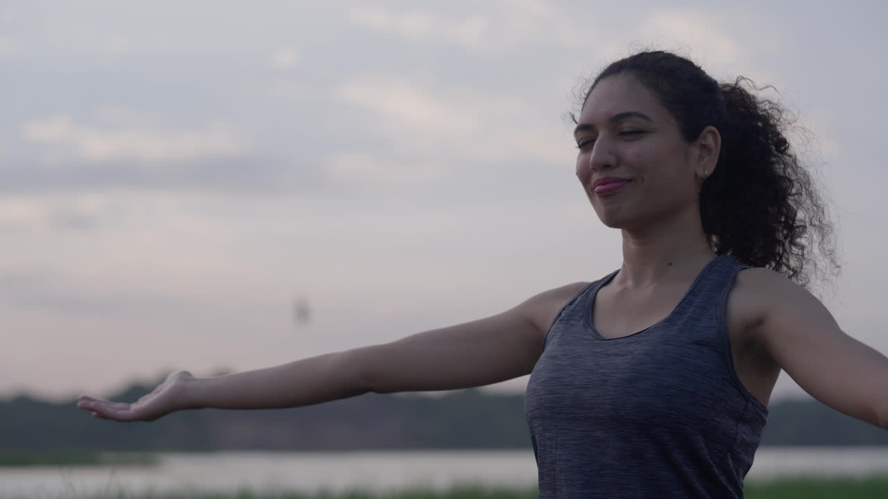 Primo piano di una donna sud-asiatica che allunga le braccia con grazia durante una tranquilla sessione di yoga all'alba in riva al lago