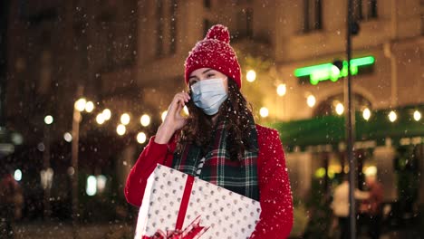 retrato de una joven hermosa y feliz hablando por teléfono inteligente y sosteniendo un regalo en la calle mientras nieva en navidad