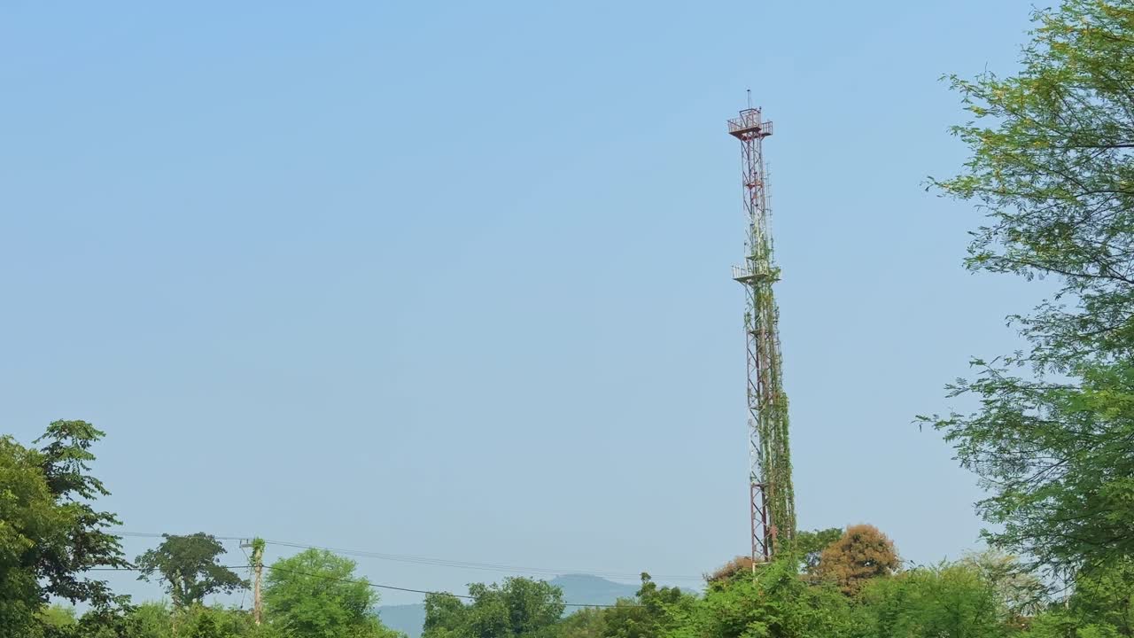 A wide static shot of a tall telecom tower surrounded by greenery and clear blue sky in a rural area