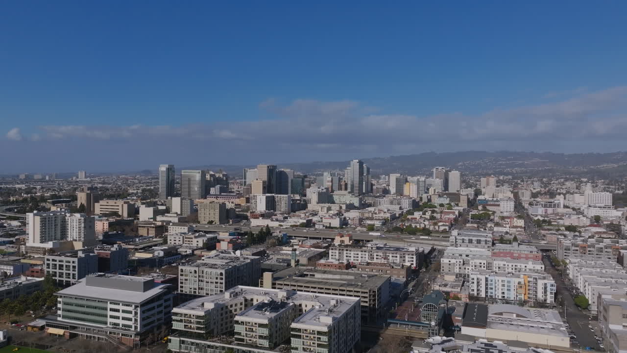 Aerial footage of downtown Oakland, California, showcasing its mix of modern high-rises, dense urban neighborhoods, and a backdrop of rolling hills under a clear blue sky.