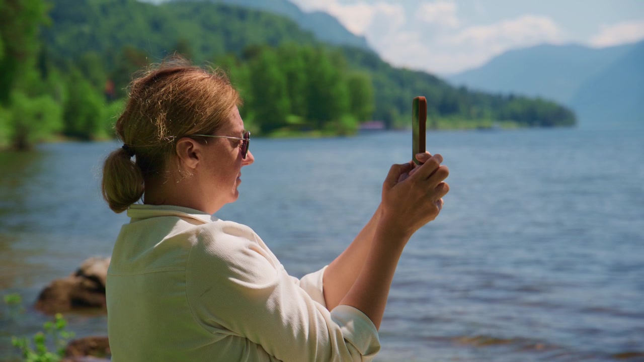 Woman stands by calm lake, phone in hand, capturing scenic view. She wears sunglasses, white shirt, hair in neat bun. Focus on peaceful moment, nature's beauty reflected in her calm posture