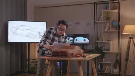 hombre asiático diseñador de automóviles comparando su diseño con el diseño de automóviles papeles de boceto mientras trabajaba en la escultura de arcilla de automóviles en el estudio