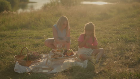 mother and daughter sitting together outdoors having picnic on sunny day, mother using smartphone while daughter snacks on fruit, enjoying time together on picnic mat with variety of snack