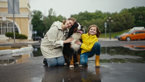 retrato de una feliz mujer rubia sentada con su hija adolescente y un perro negro mientras camina en el parque después de la lluvia