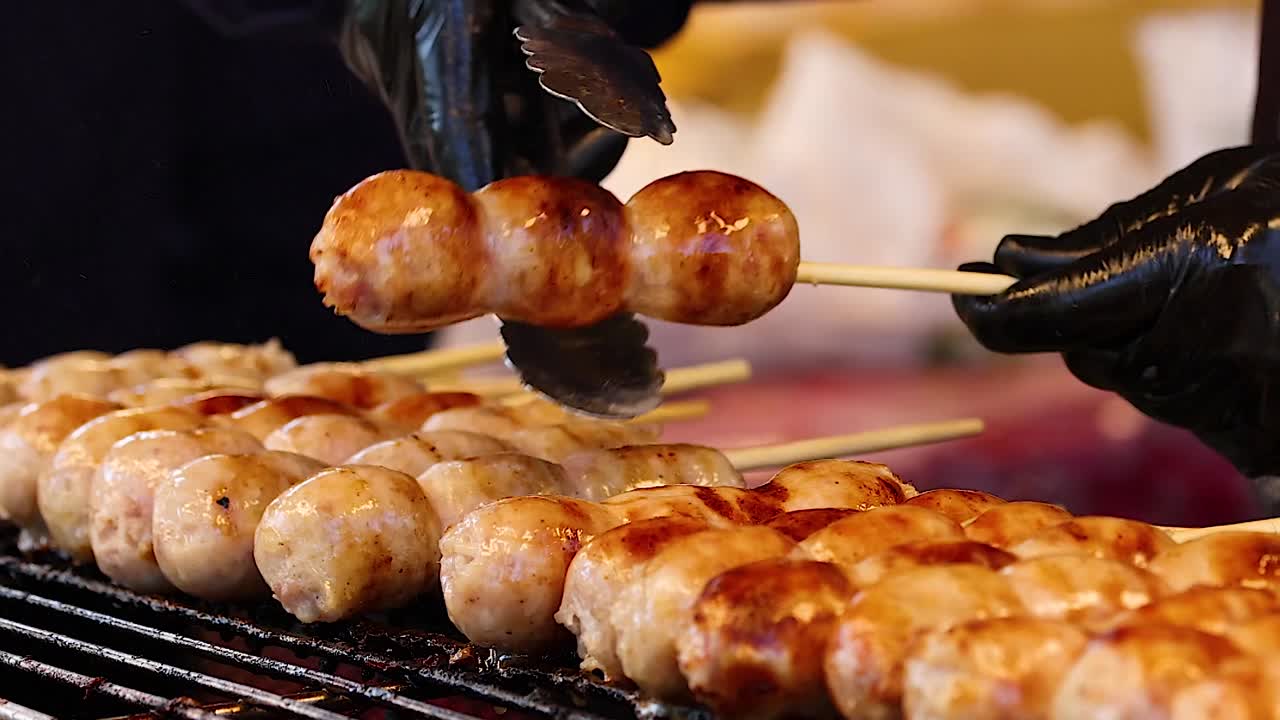 屋台の食べ物屋が金属製のトングを使って、串に刺した焼きソーセージをひっくり返しています。