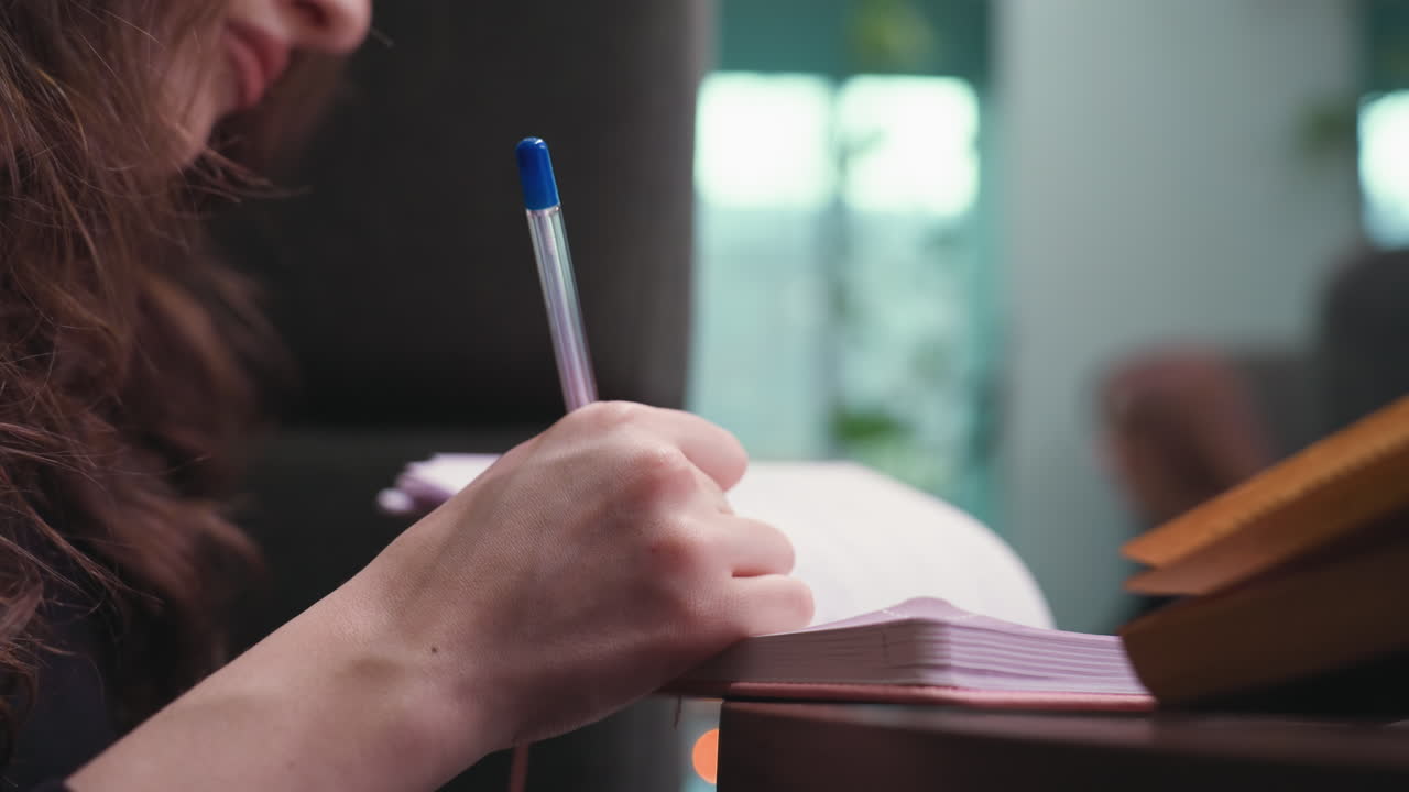 Close up of woman hand holding blue pen while writing in notebook on wooden table, with soft light and blurred indoor background creating peaceful, focused study or journaling atmosphere