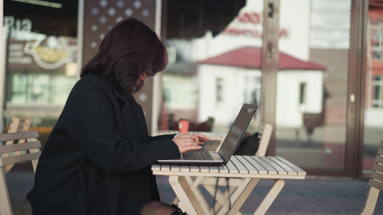 Side view of lady focused on laptop screen while seated outdoors in urban environment under natural light, scene features modern furniture, a blurred background of building