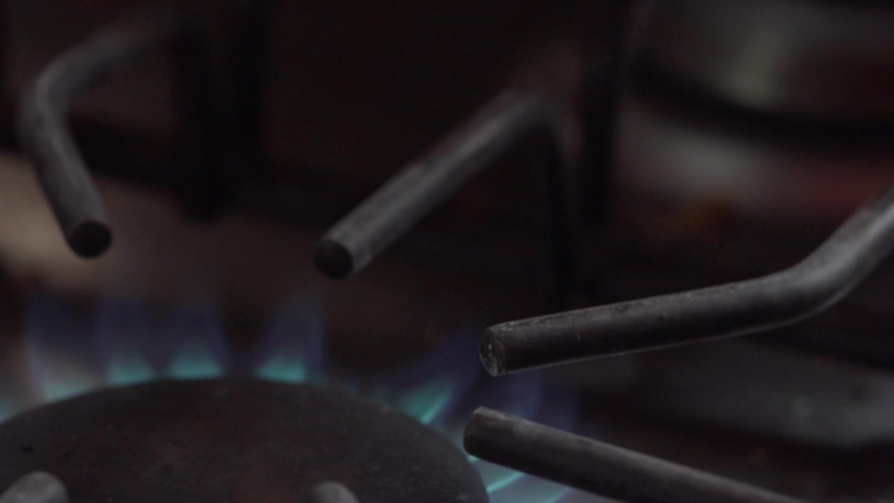Closeup of a blue gas flame burning beneath a metal stove burner during cooking. Low flames flicker around the burner, highlighting heat, combustion and kitchen appliance details