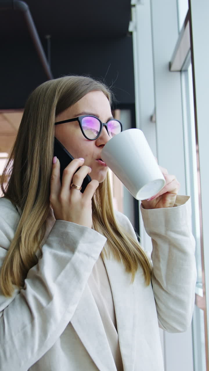 Phone talk and drinking tea during lunch break in the office. Young woman standing in the spacious office in front of large window. Vertical video