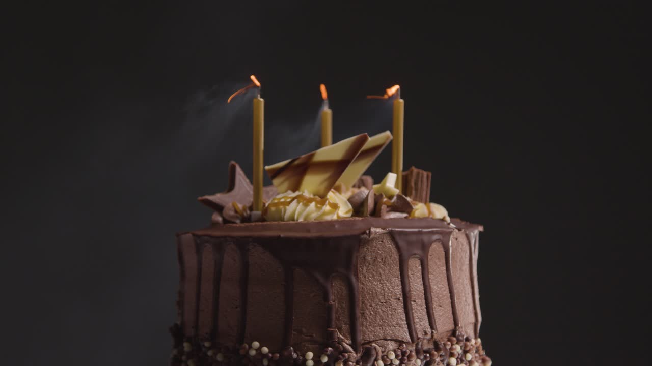 Close Up Studio Shot Of Decorated Chocolate Birthday Celebration Cake With Candles Being Blown Out Against Black Background