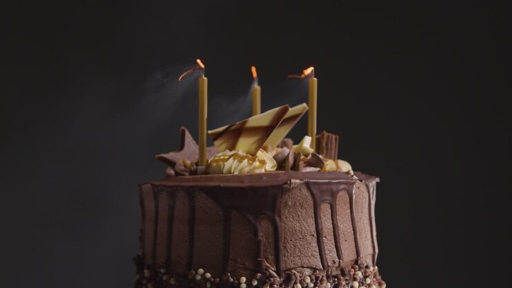 Close Up Studio Shot Of Decorated Chocolate Birthday Celebration Cake With Candles Being Blown Out Against Black Background