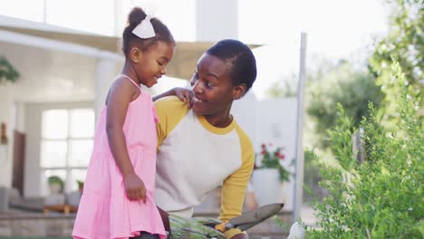 Happy-african-american-mother-and-daughter-gardening-and-talking,-mother-holding-trowel