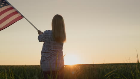 young american woman waving the us flag against the backdrop of a field of wheat where the sun rises